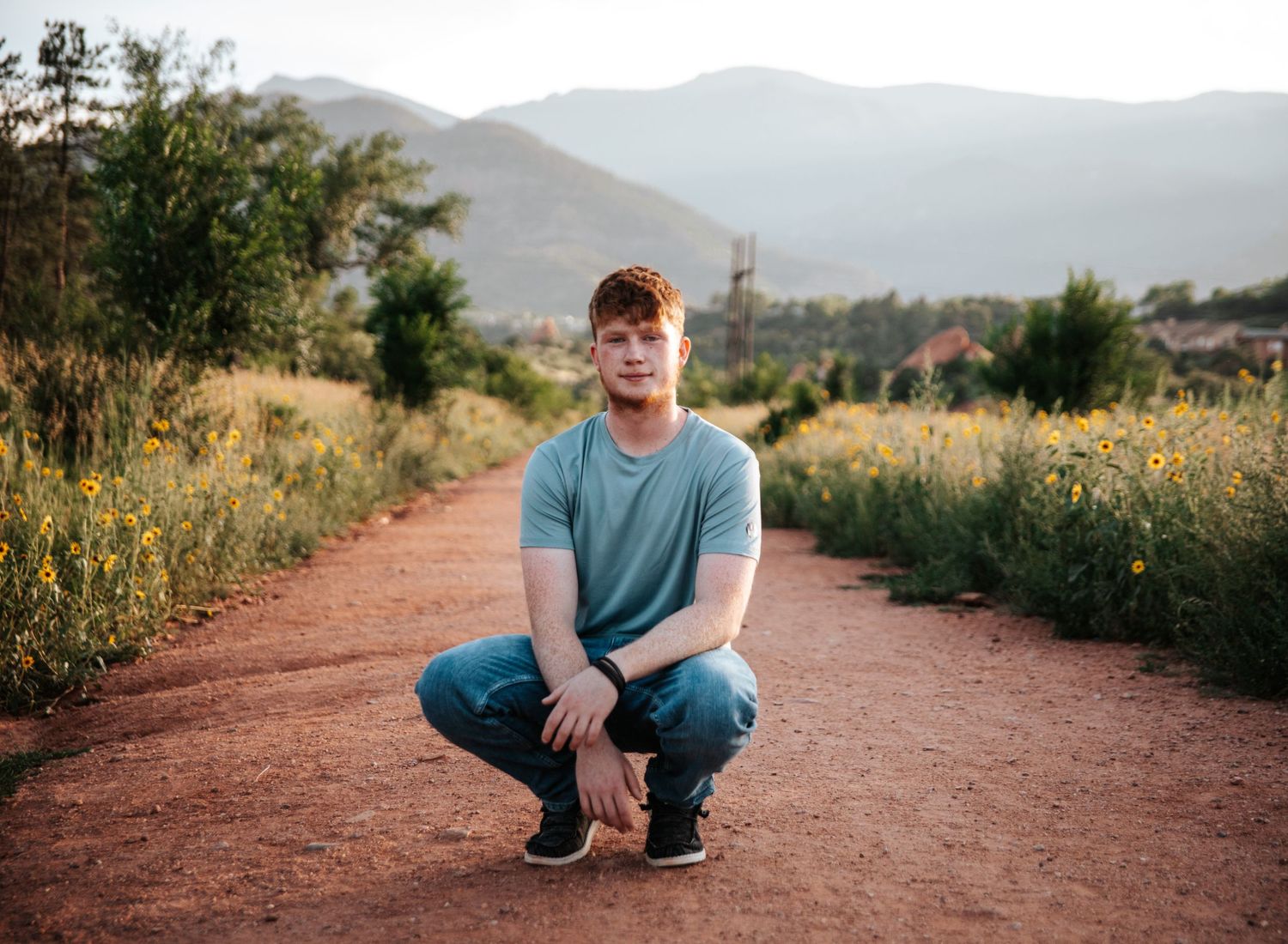 Person crouching on a red dirt trail with mountains visible in the background.