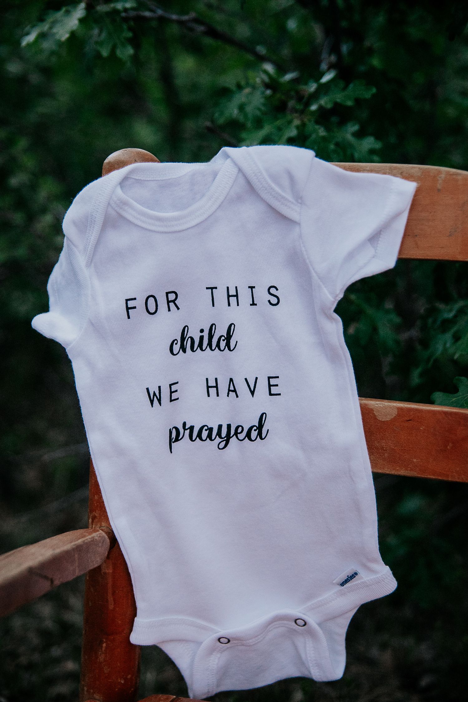 White baby onesies with text displayed on wooden chairs against green foliage.
