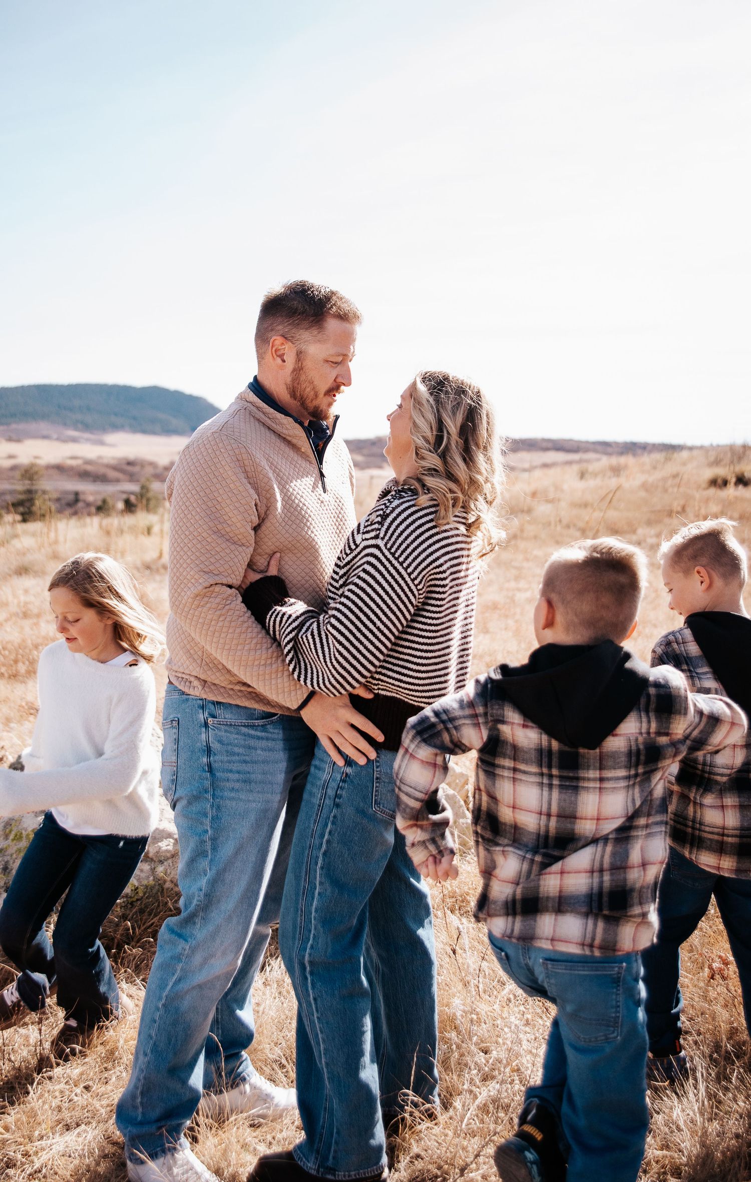 A couple shares an intimate moment outdoors while children play in the background on a sunny hillside.