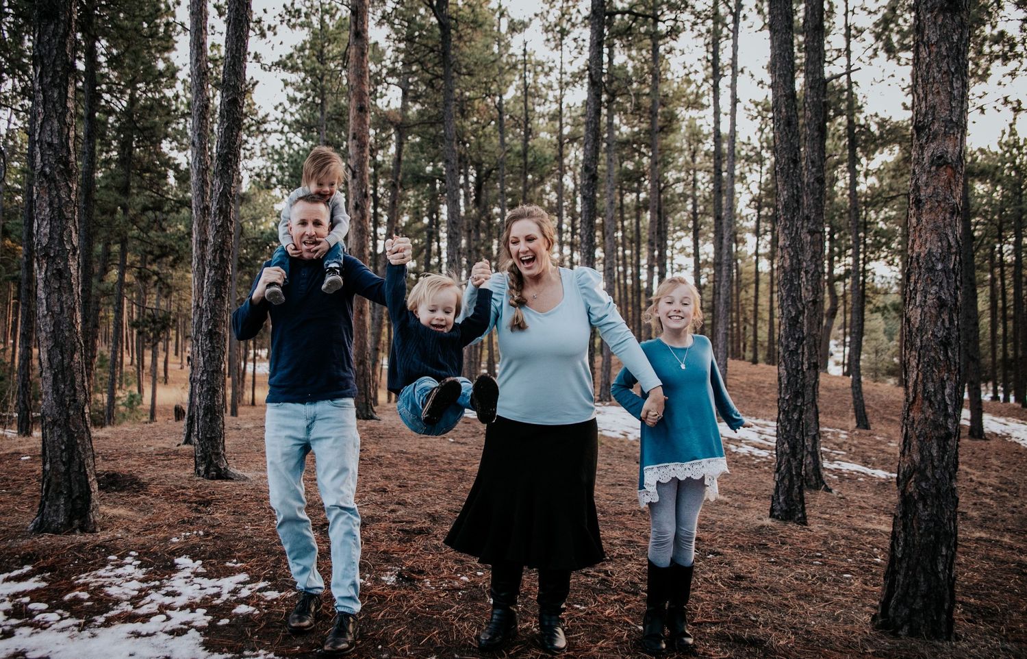 Playful family photo of four people jumping and running through snowy forest.