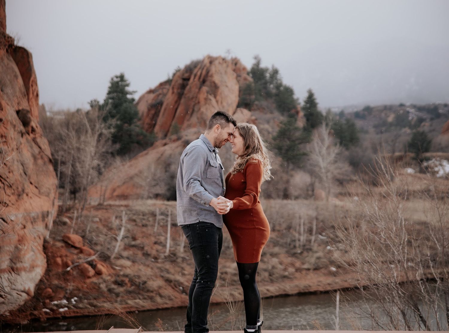 A couple poses for maternity photos along a scenic lakeside trail with red rock formations.