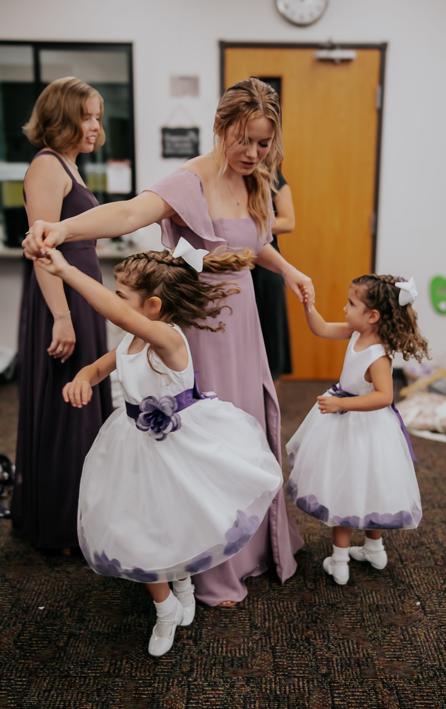 Young flower girls in white and purple dresses twirling on carpeted floor.
