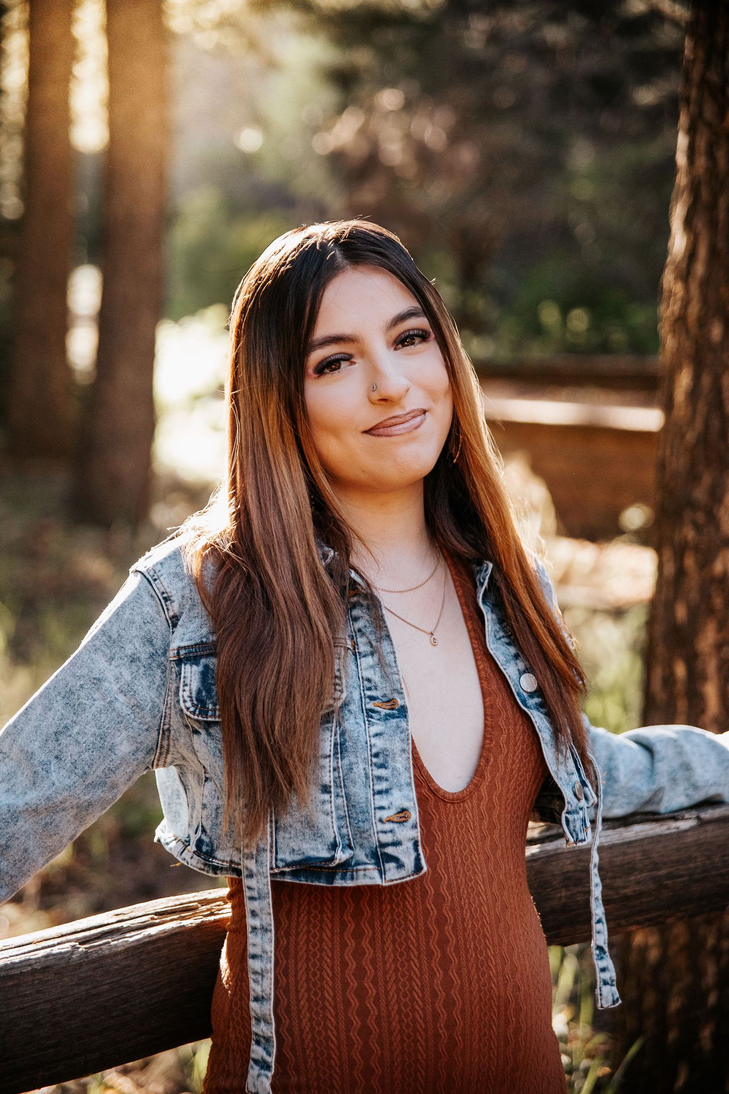 Portrait in sunlit forest setting leaning on wooden fence wearing denim and brown outfit.
