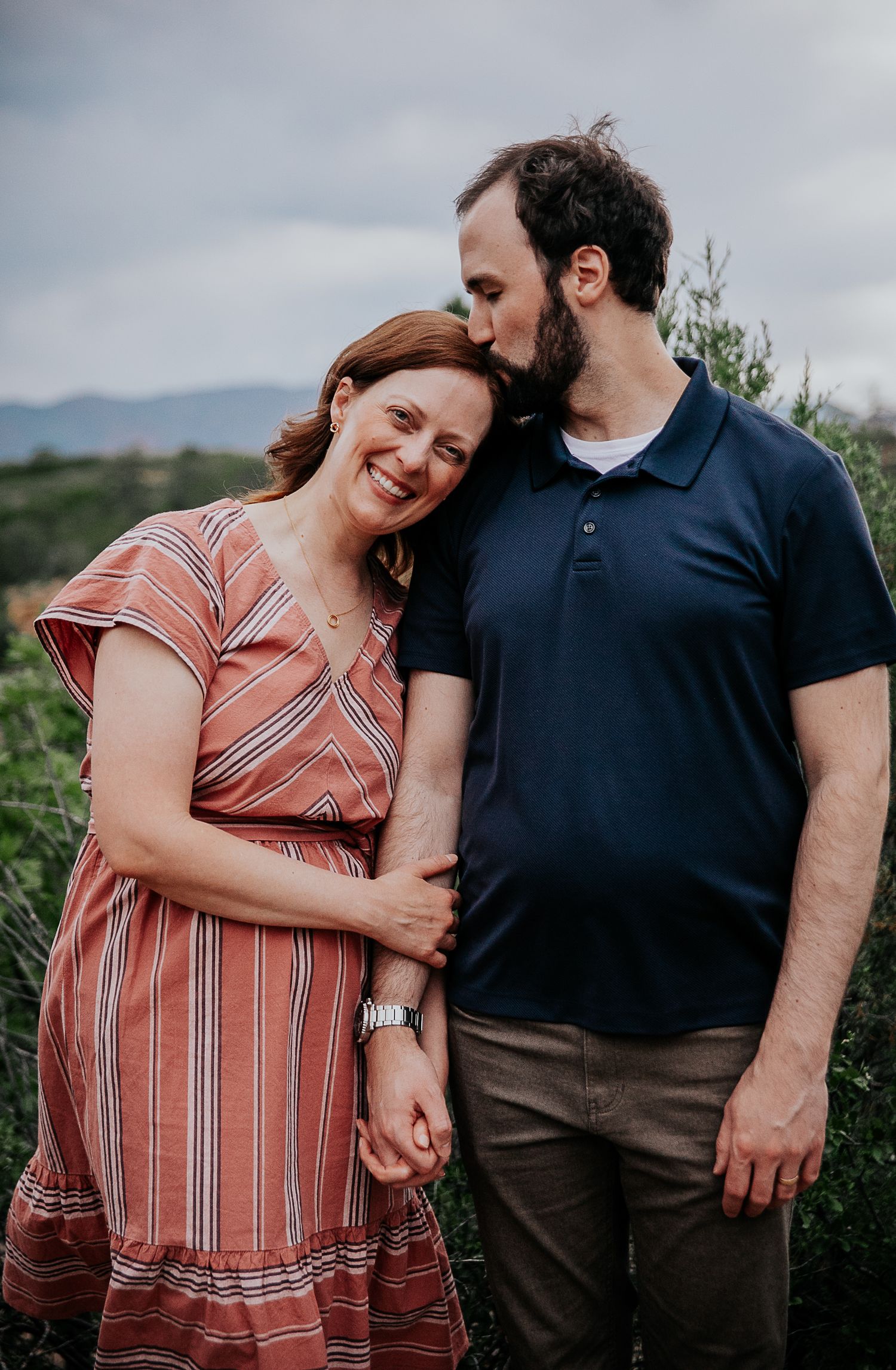 A couple embraces on a hillside, with one wearing a coral striped dress and the other in navy.