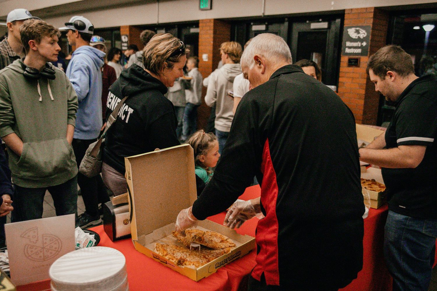 A crowded indoor event with people gathering around tables with food and refreshments.