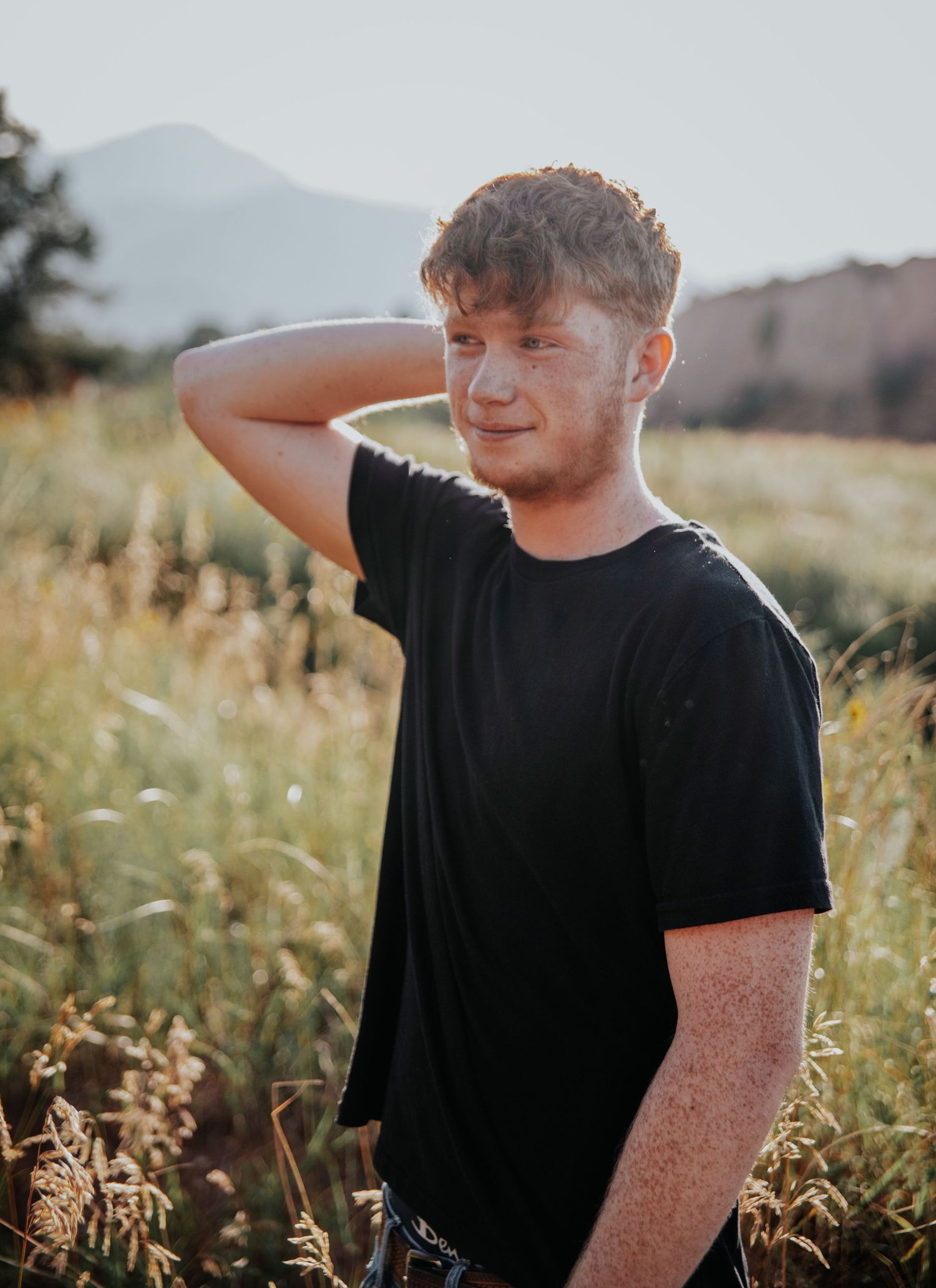 Person in black t-shirt poses in sunlit meadow with mountains visible in background.