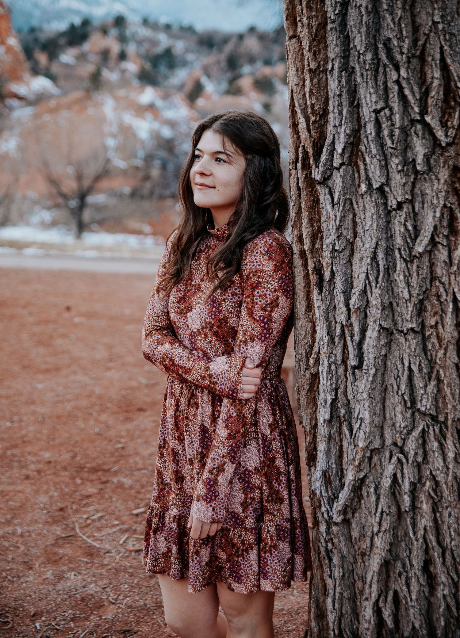 A woman in a vintage-style dress leans against a large tree trunk with mountains in the background.