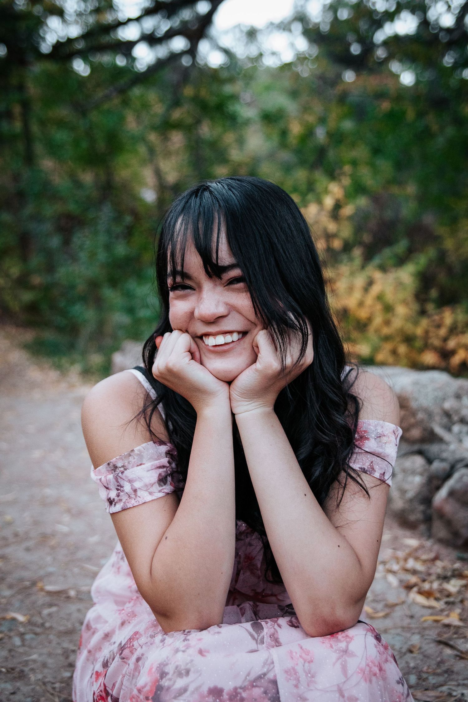 A joyful person in a pink floral dress laughs while sitting outdoors in a forest setting.