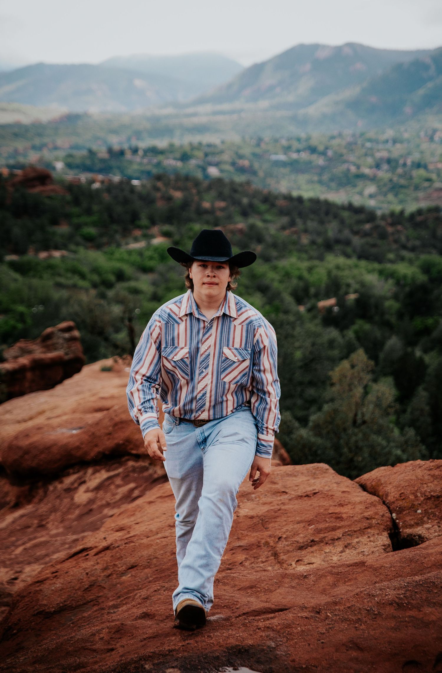 Person in cowboy hat and striped shirt sits atop red rocks with mountain vista in background at Garden of the Gods in Colorado Springs