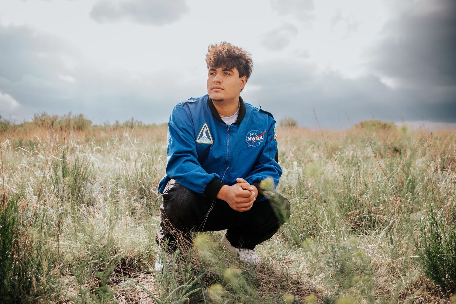 High school senior in blue NASA flight jacket crouching in a grassy field on an overcast day at Palmer Park in Colorado Springs