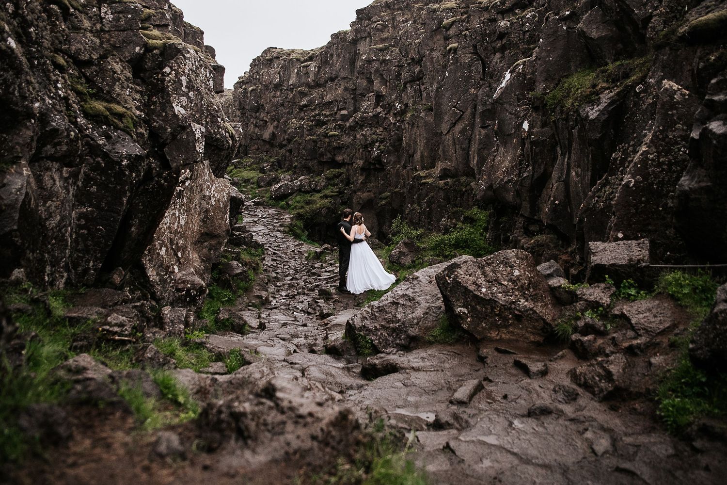 A couple poses dramatically between rocky cliffs in their wedding attire.