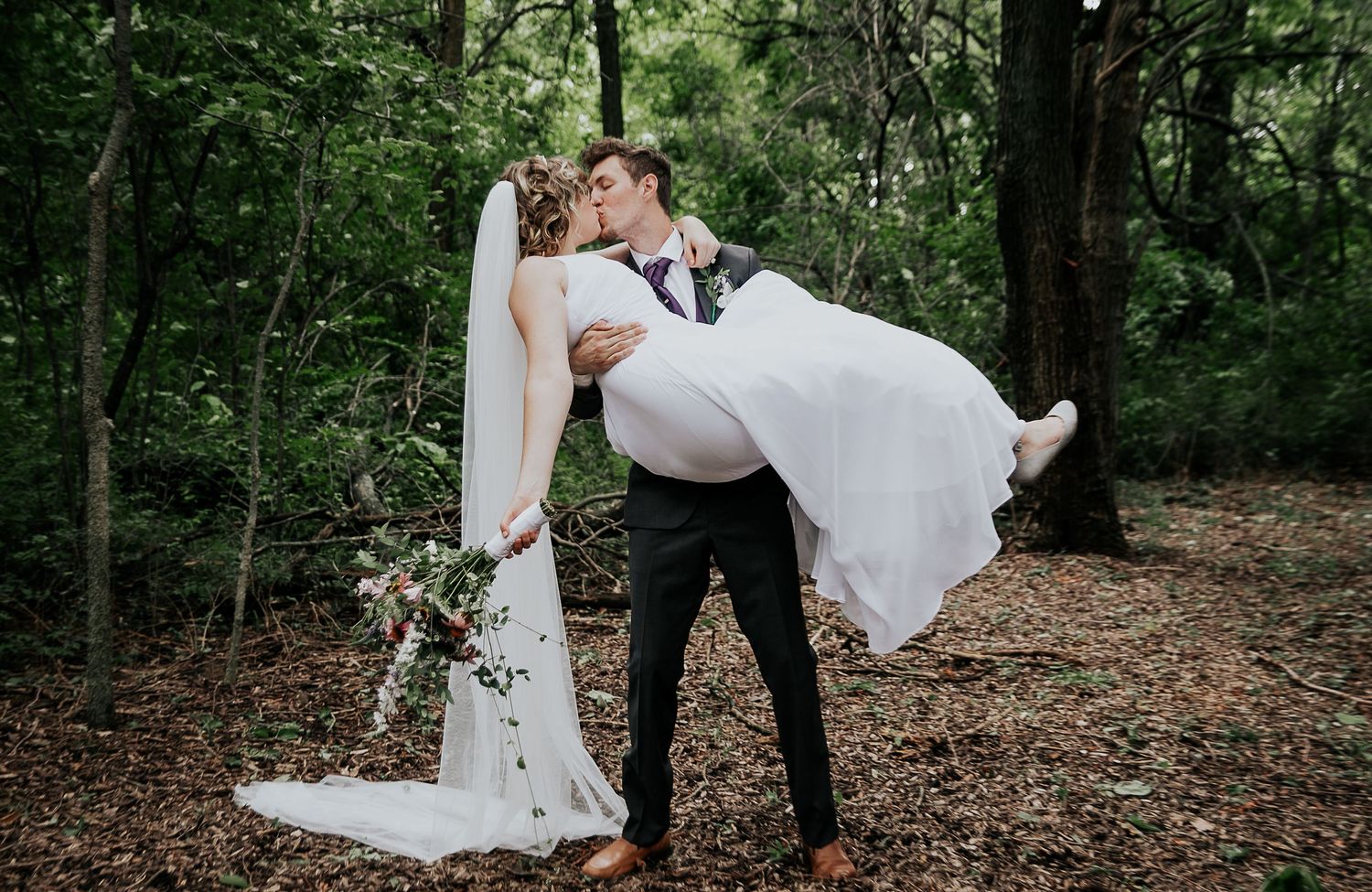 A romantic wedding couple embraces in a wooded forest setting, the groom lifting the bride in her flowing white dress.
