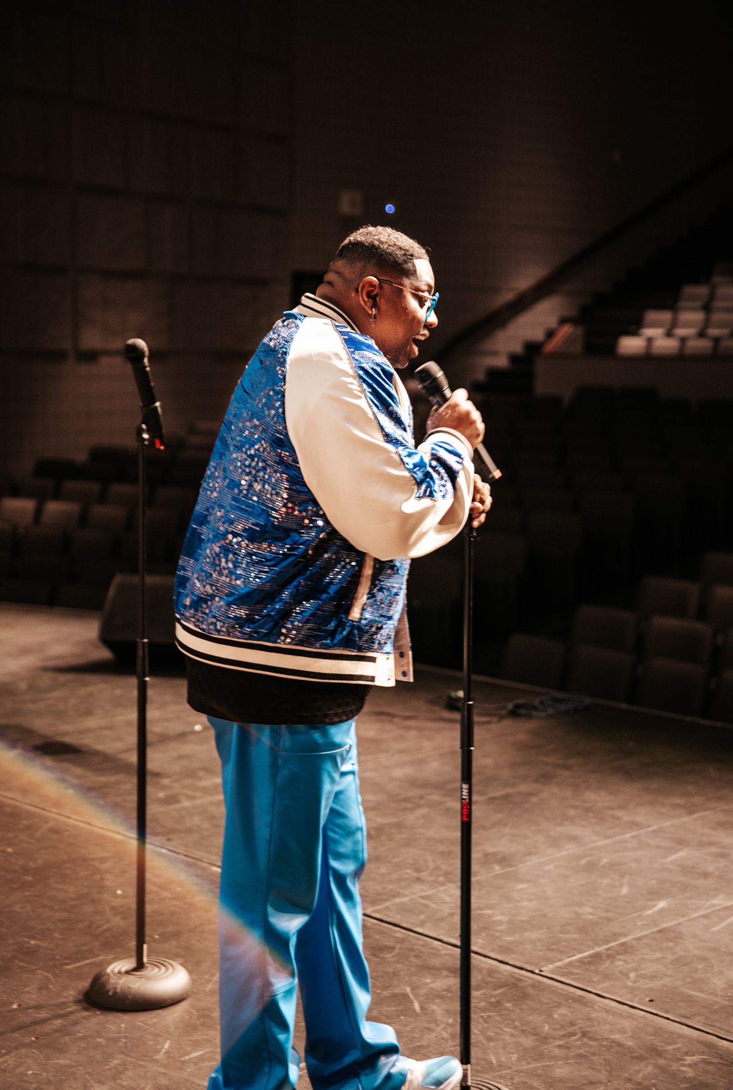 Performer in a bright blue and white patterned outfit sings on a dimly lit stage.
