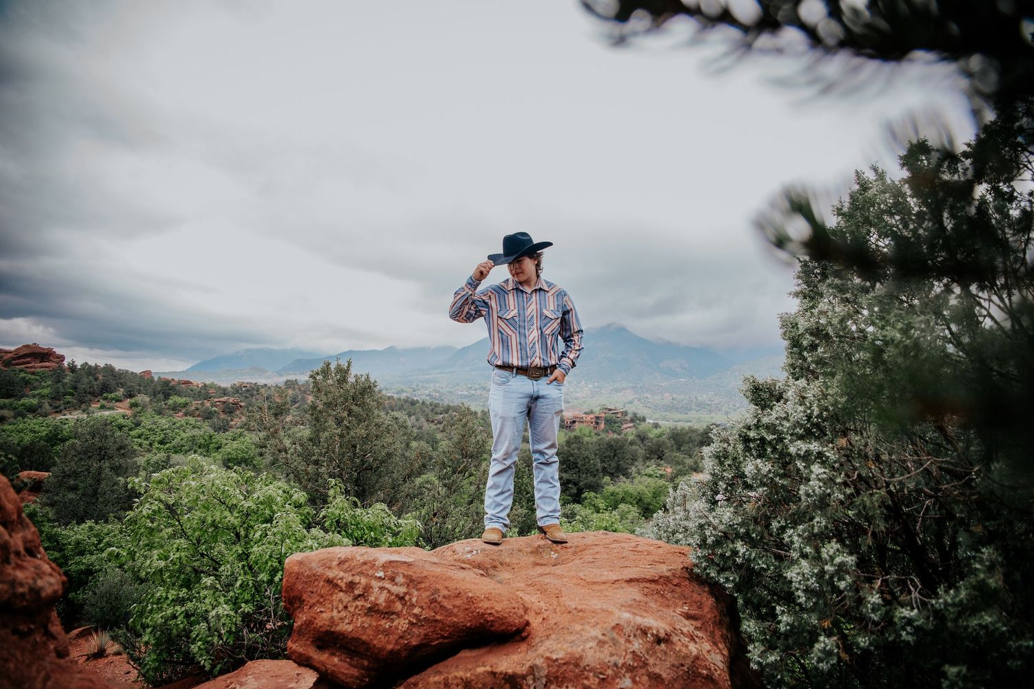 A high school senior in western attire touches their hat brim while posing on red rocks at Garden of the Gods in Colorado Springs