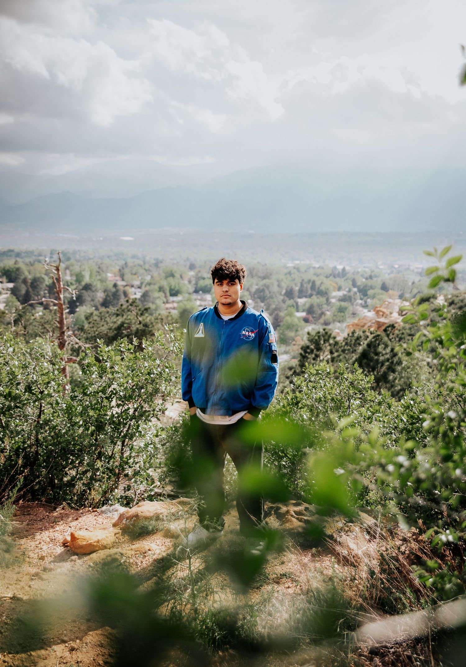 High school senior with blue NASA jacket visible through foliage with cityscape view in background at Palmer Park in Colorado Springs