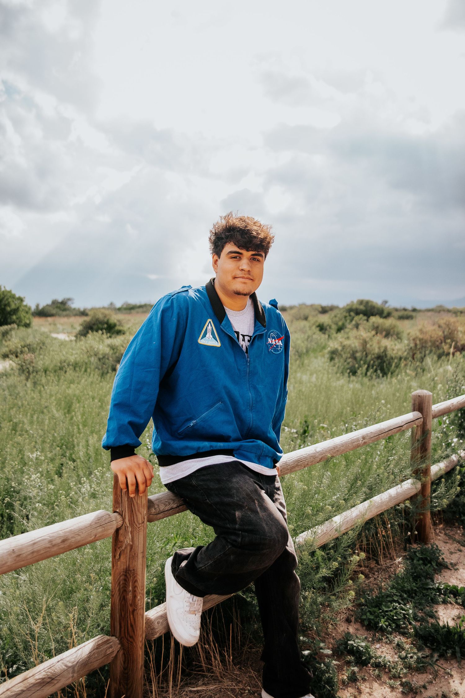 High school senior in a NASA jacket sits casually on wooden fence rails with cloudy sky in background at Palmer Park in Colorado Springs