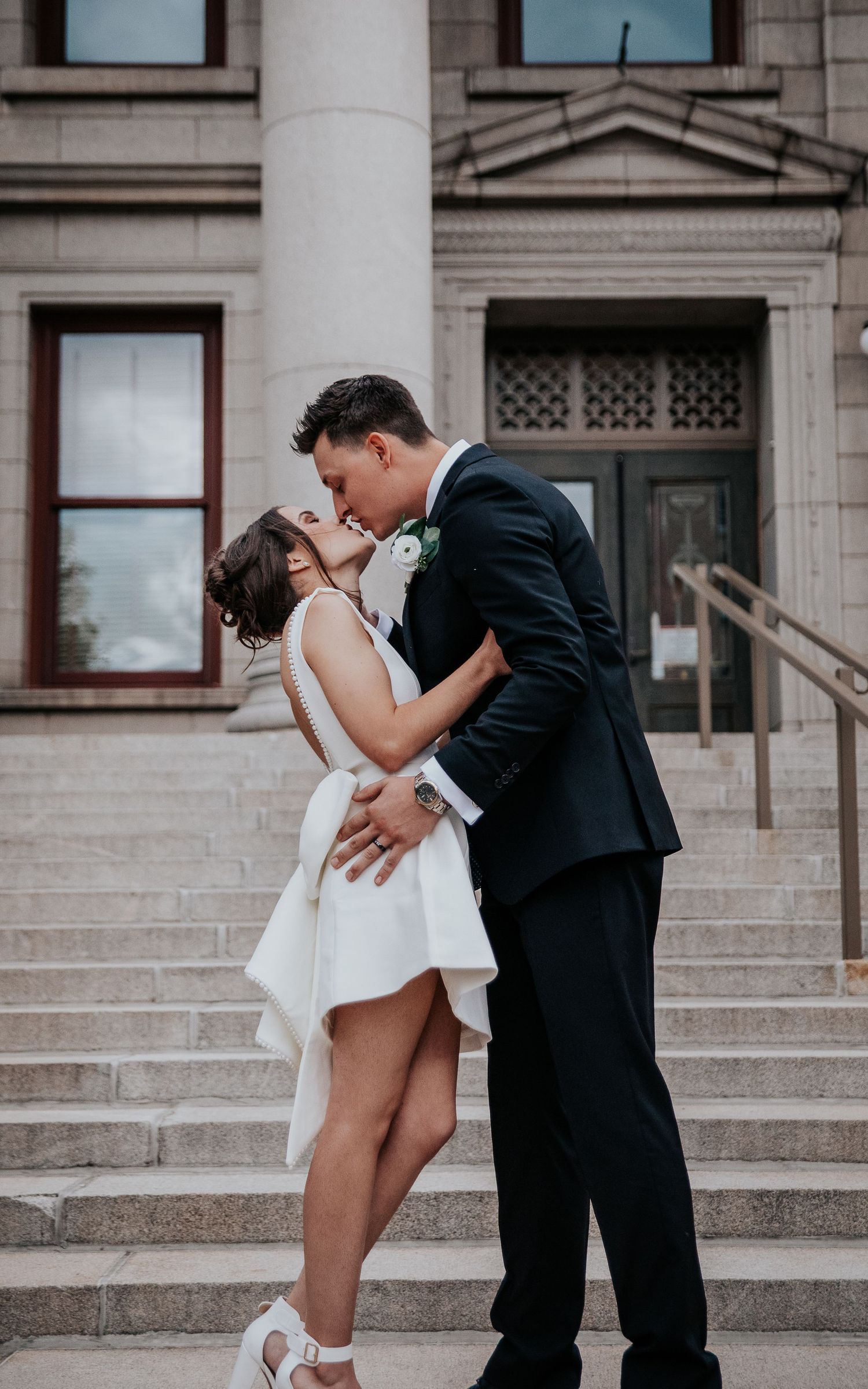 A romantic couple in formal attire share an intimate moment on stone steps of a historic building.