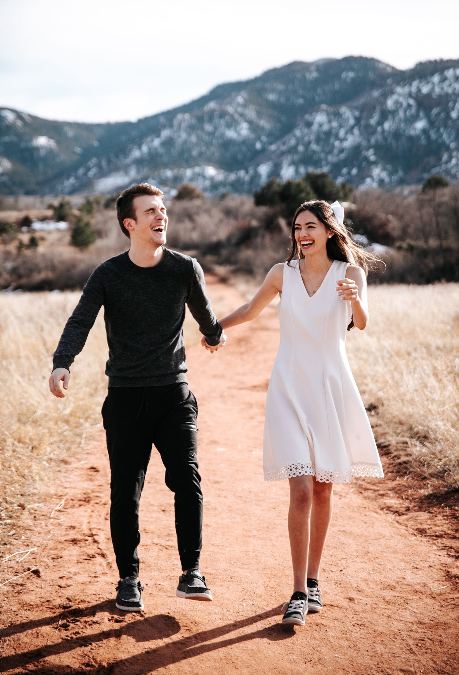A couple joyfully runs down a dirt trail together with mountains in the background.