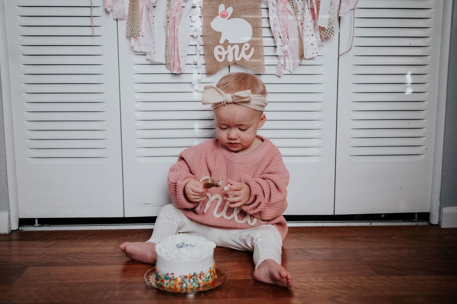 First birthday setup with white shutters, pink decorations, and small white cake on wooden floor.