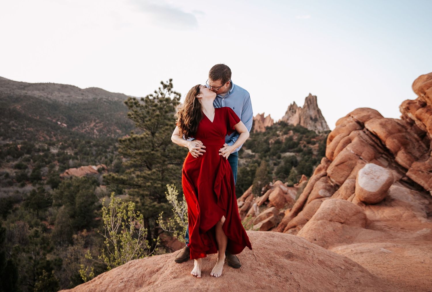 Couple in red and blue outfits embracing on red rocks with mountain vista behind them.