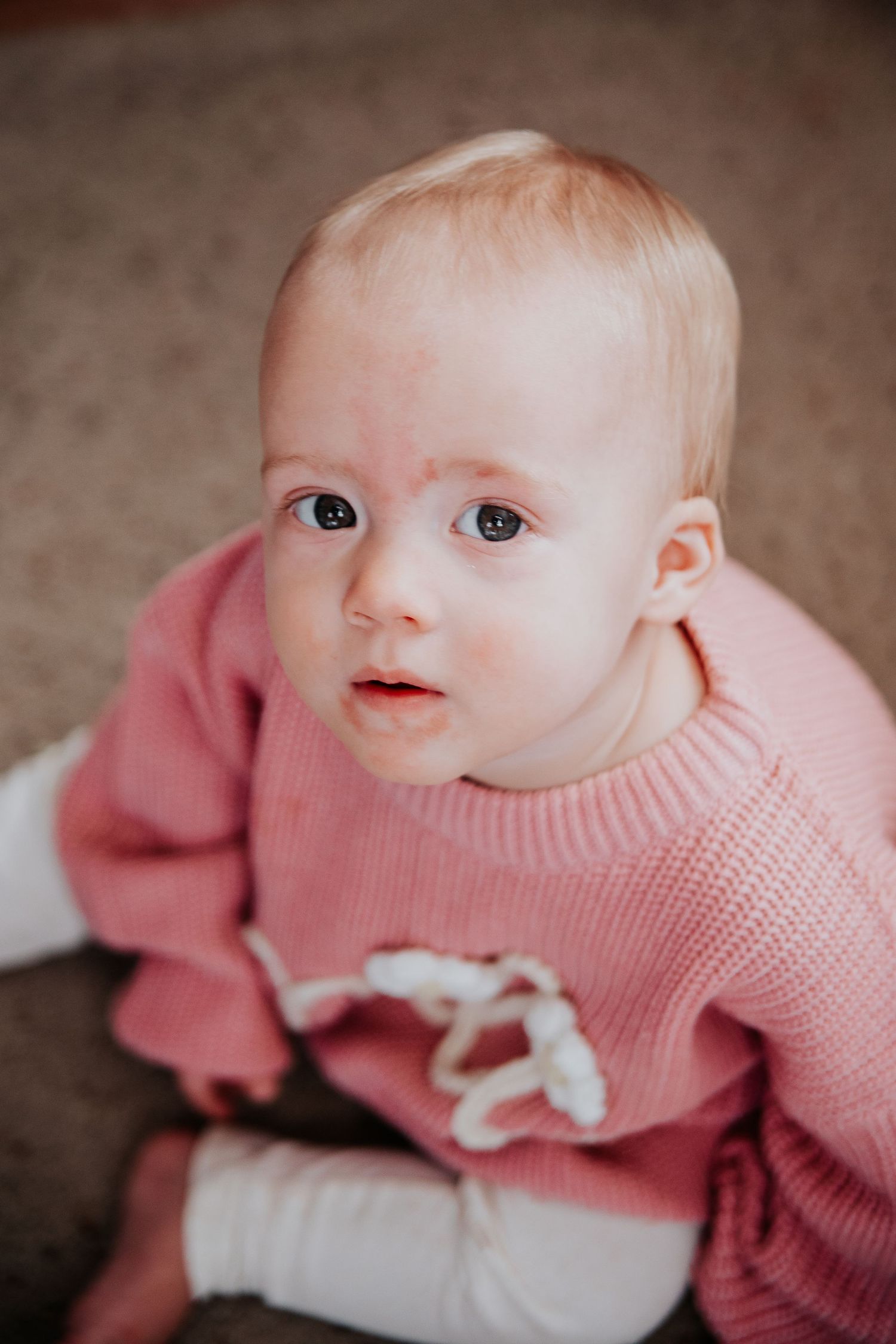 Baby wearing pink knit sweater sitting on carpet looking up at camera.