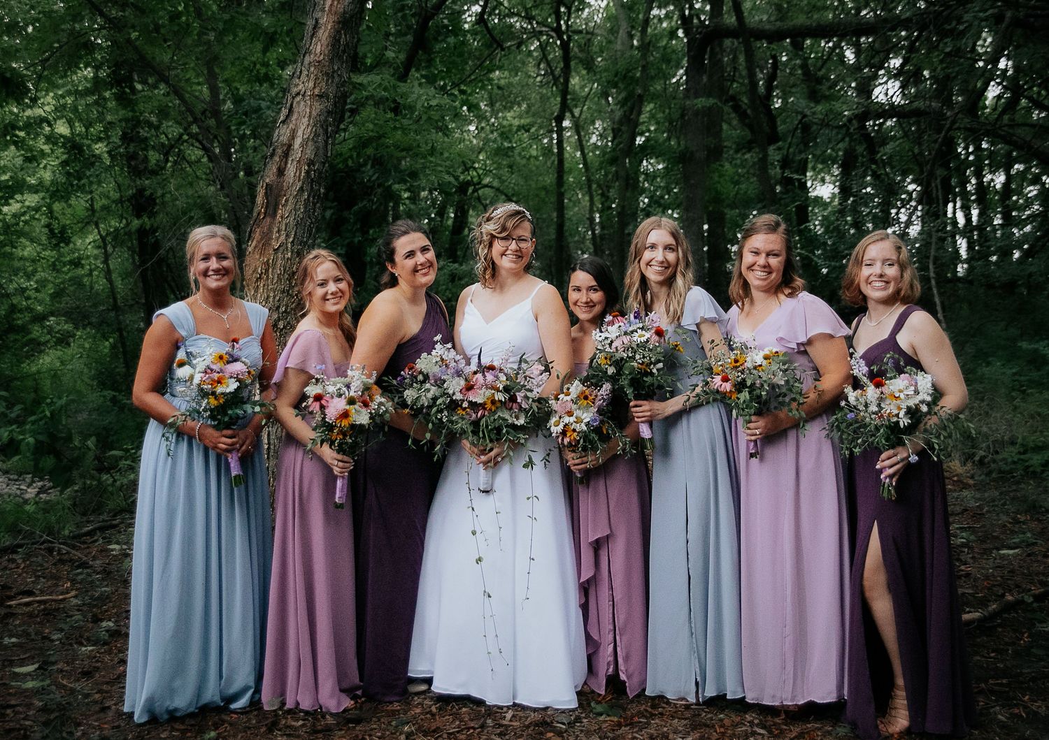 Wedding party poses in a woodland setting wearing dusty blue and mauve bridesmaids dresses with colorful bouquets.