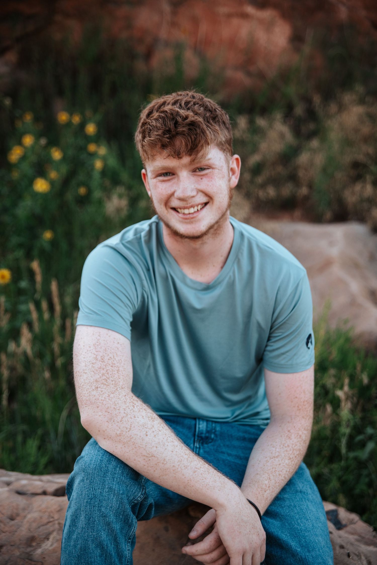 Portrait of person in light blue t-shirt sitting on rock with wildflowers in background.