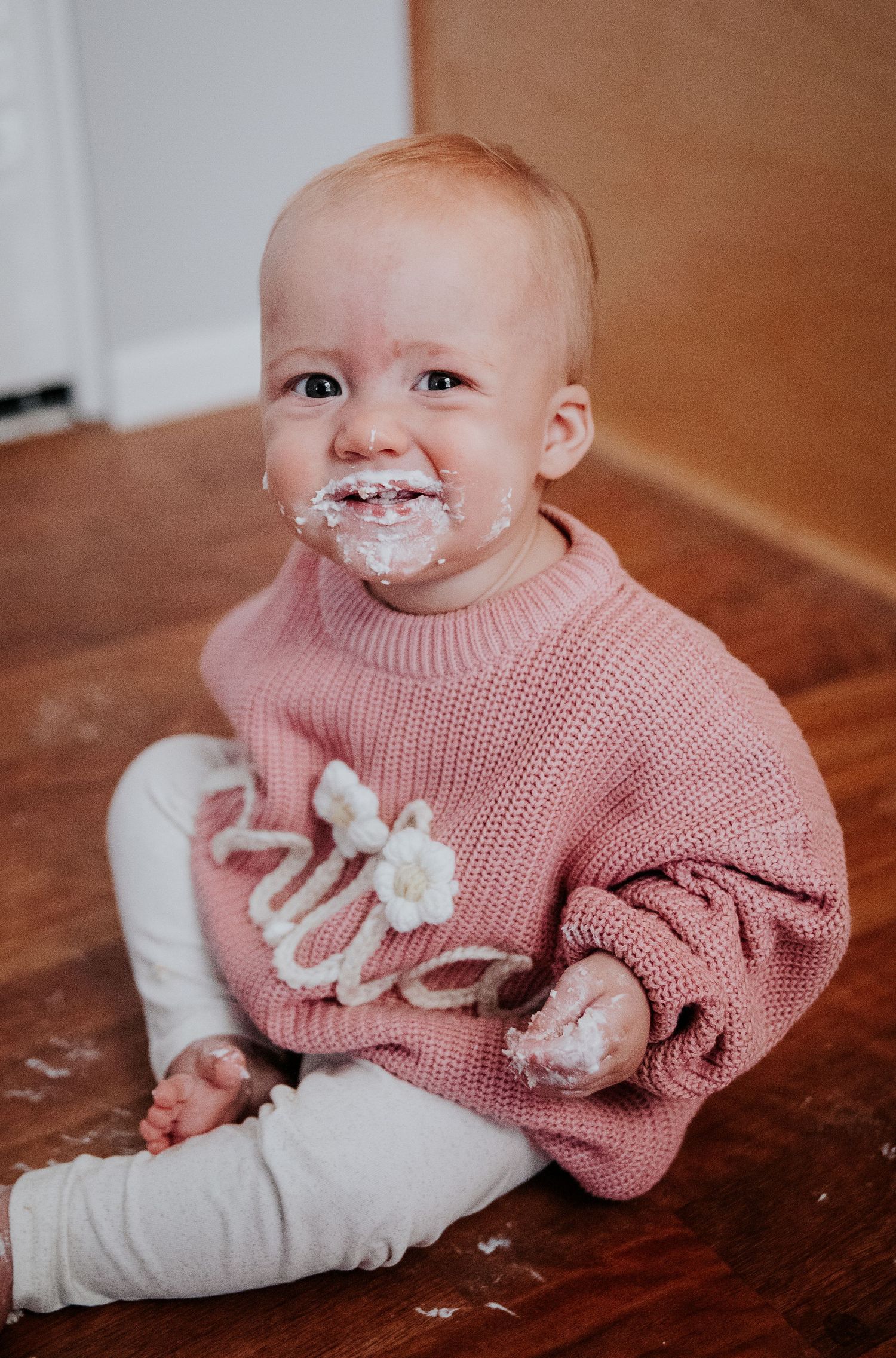 A smiling baby wearing pink sits on hardwood floor with cake frosting on their face.