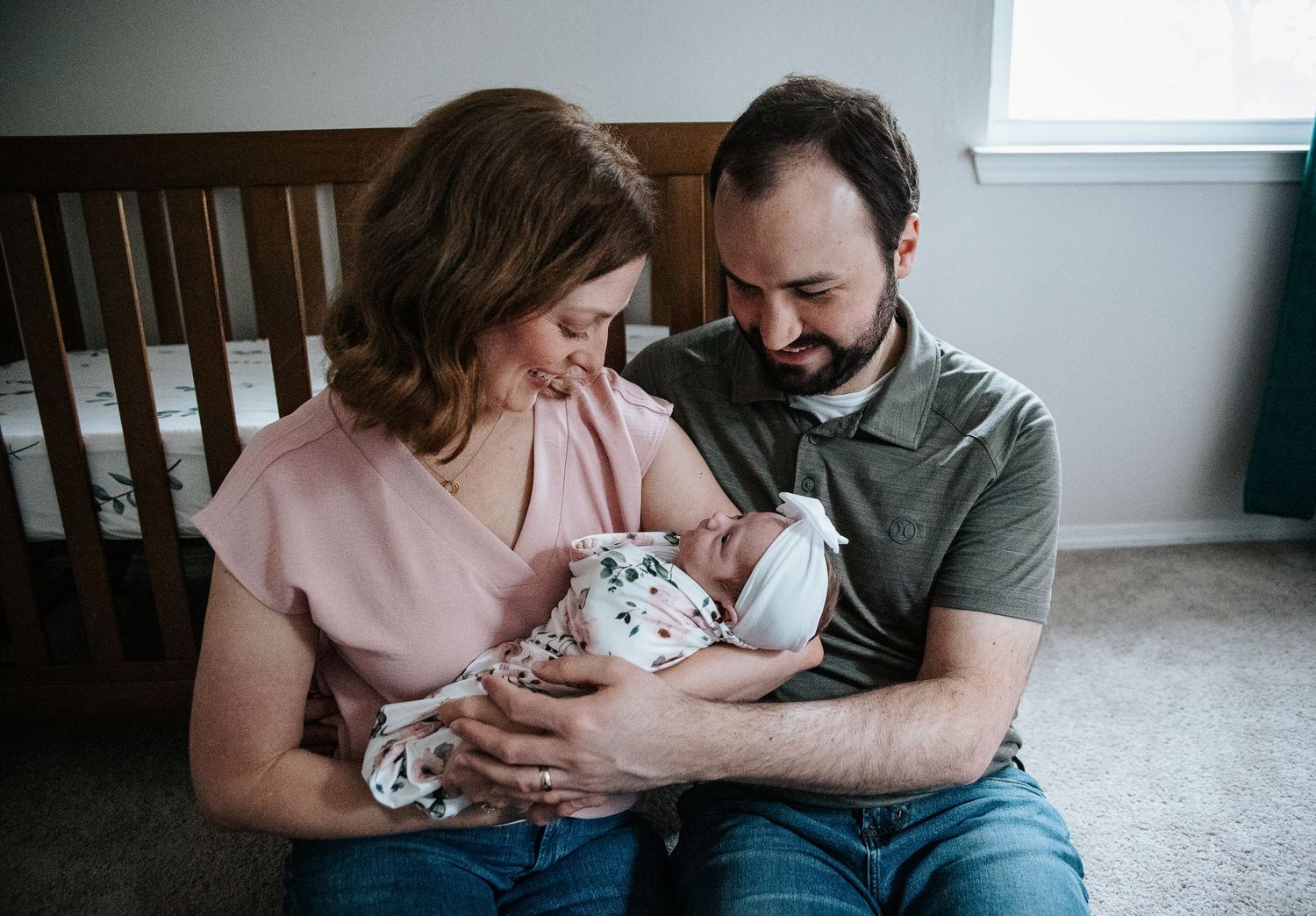 Parents sitting together on floor by wooden crib sharing intimate moment with their newborn baby.
