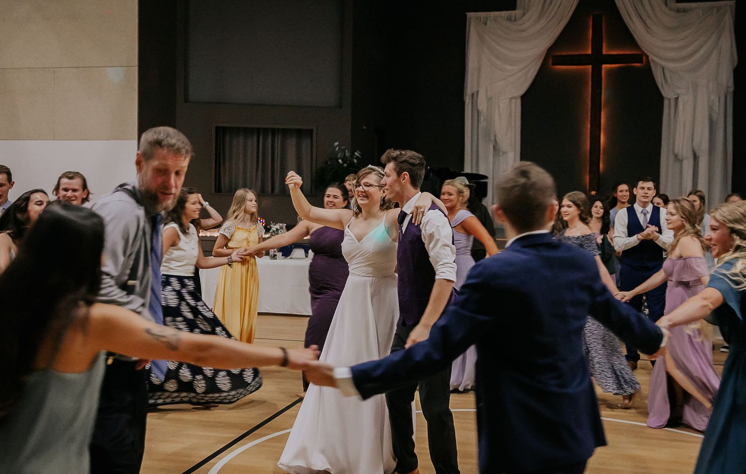 Wedding guests dancing joyfully in a reception hall with a cross visible on the back wall.