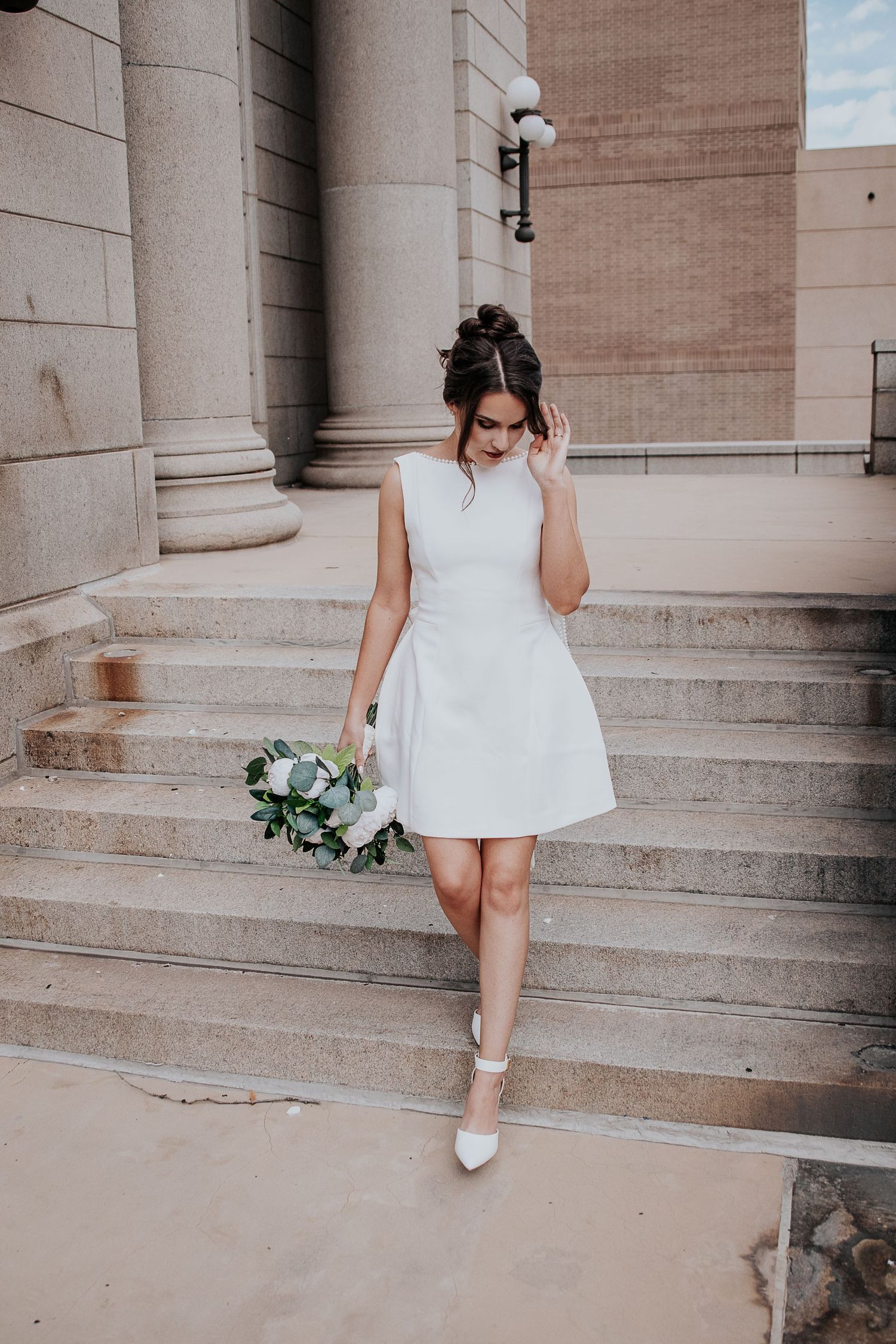 A bride in a short white dress walks down stone steps while holding a bouquet of flowers at Colorado Springs courthouse wedding.