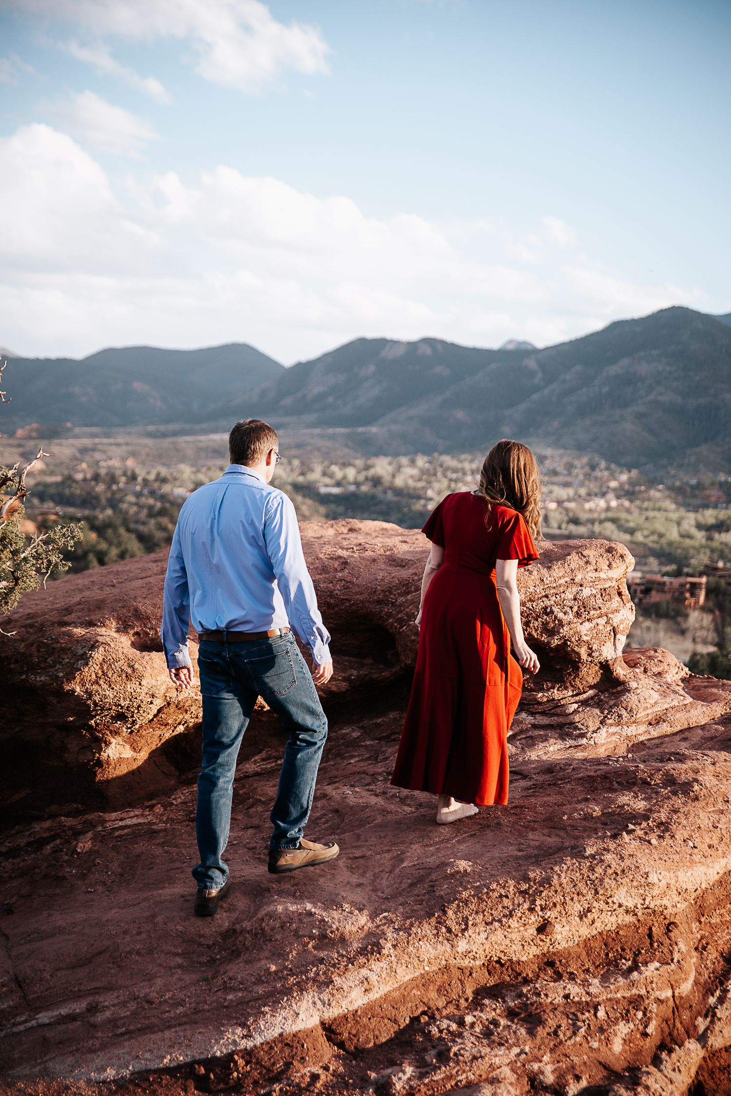 Two people walking on red rock trail overlooking mountain landscape.