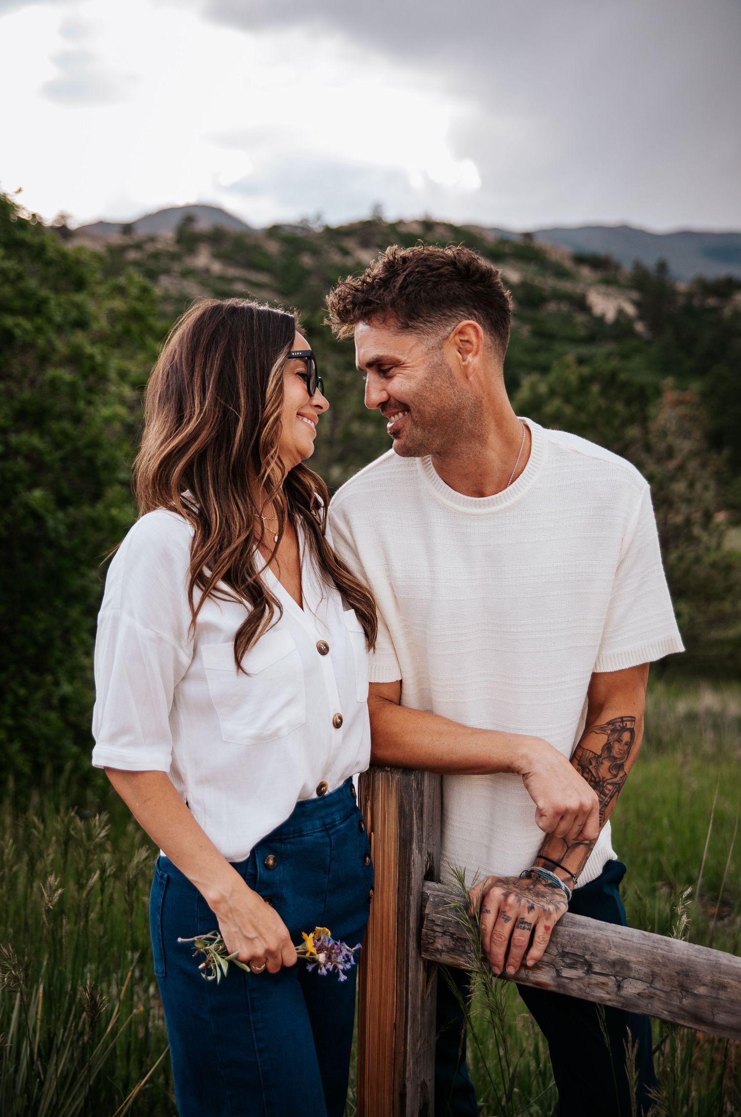 Couple in casual attire share intimate moment by wooden fence with mountain landscape behind at Ute Valley Park in Colorado Springs
