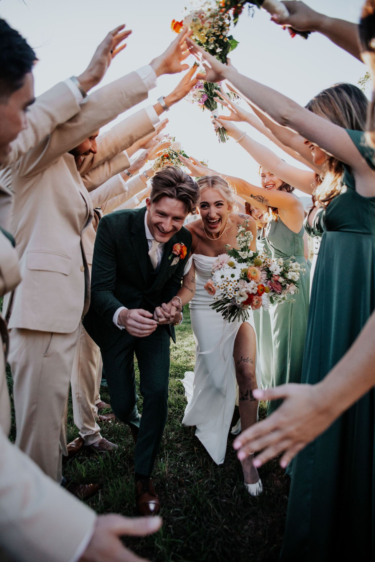 Wedding guests form a tunnel with outstretched arms as newlyweds pass underneath during outdoor celebration.