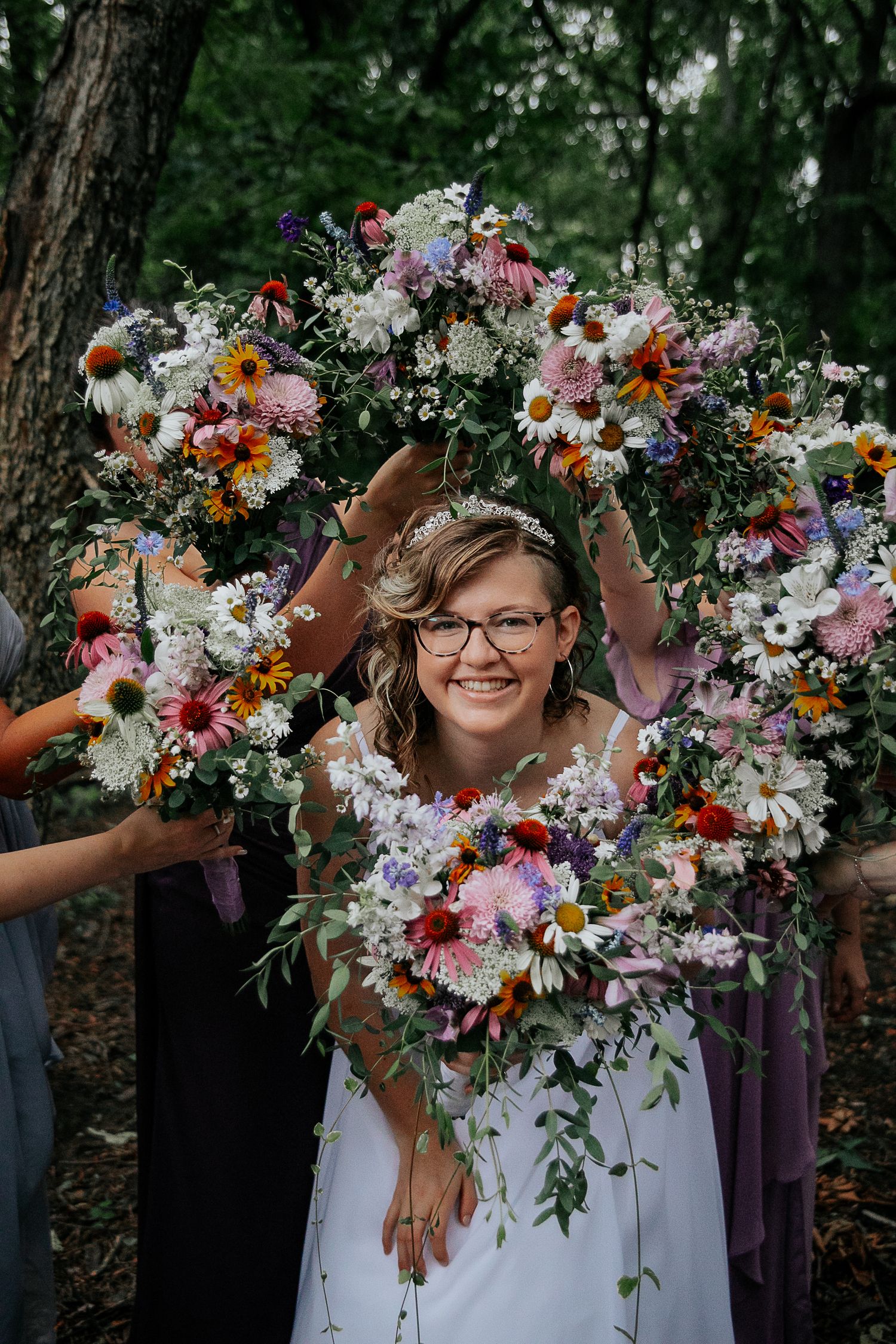 Joyful bride holds up a vibrant floral arch made of pink, orange and white wildflowers in a forest setting.