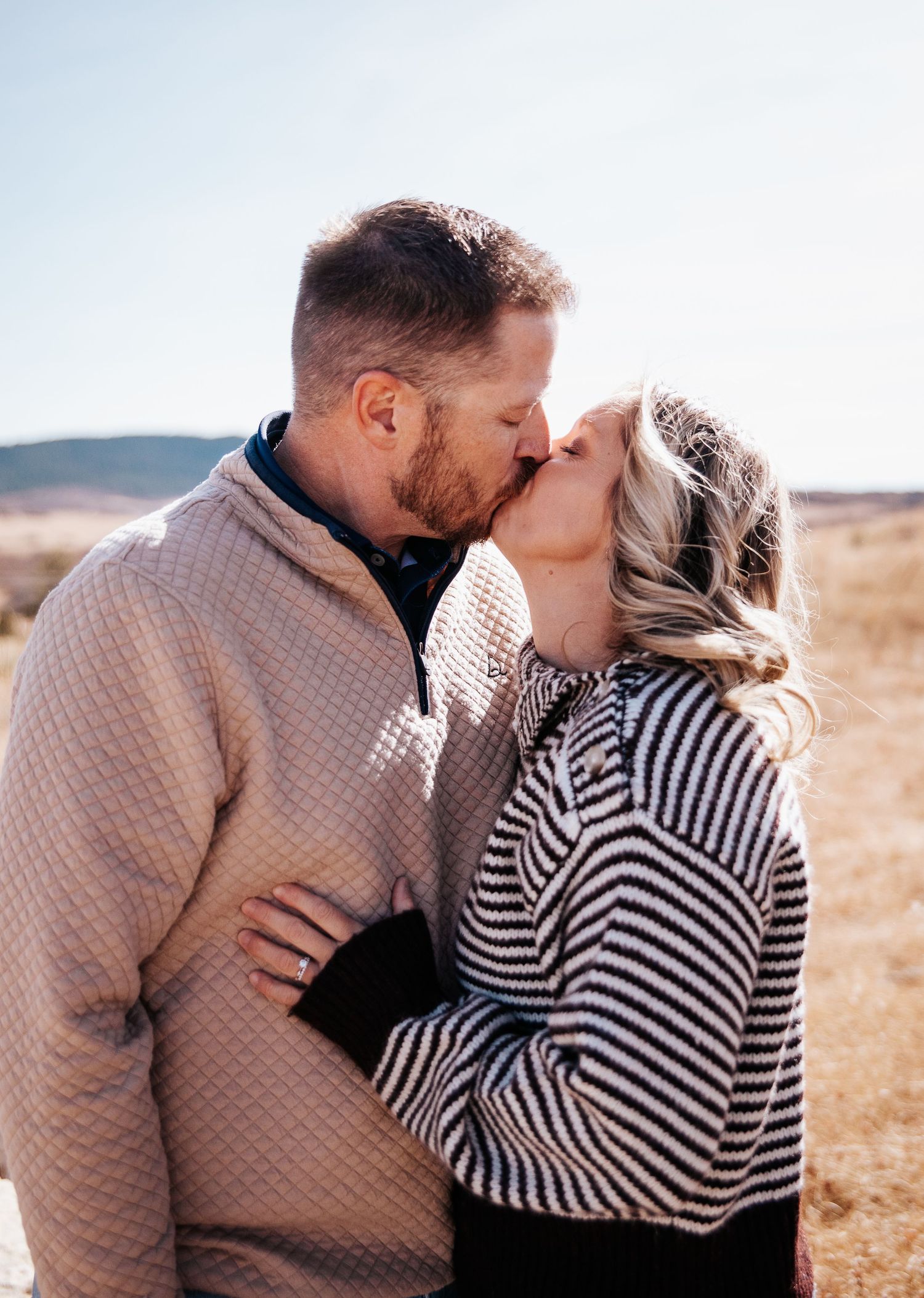 A couple in coordinating sweaters shares a romantic kiss in a rustic outdoor setting.