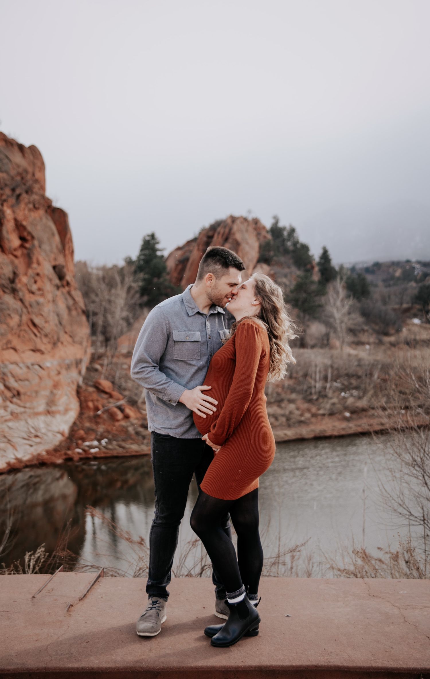 A couple shares tender moments during their maternity shoot by a lake with red rock cliffs.