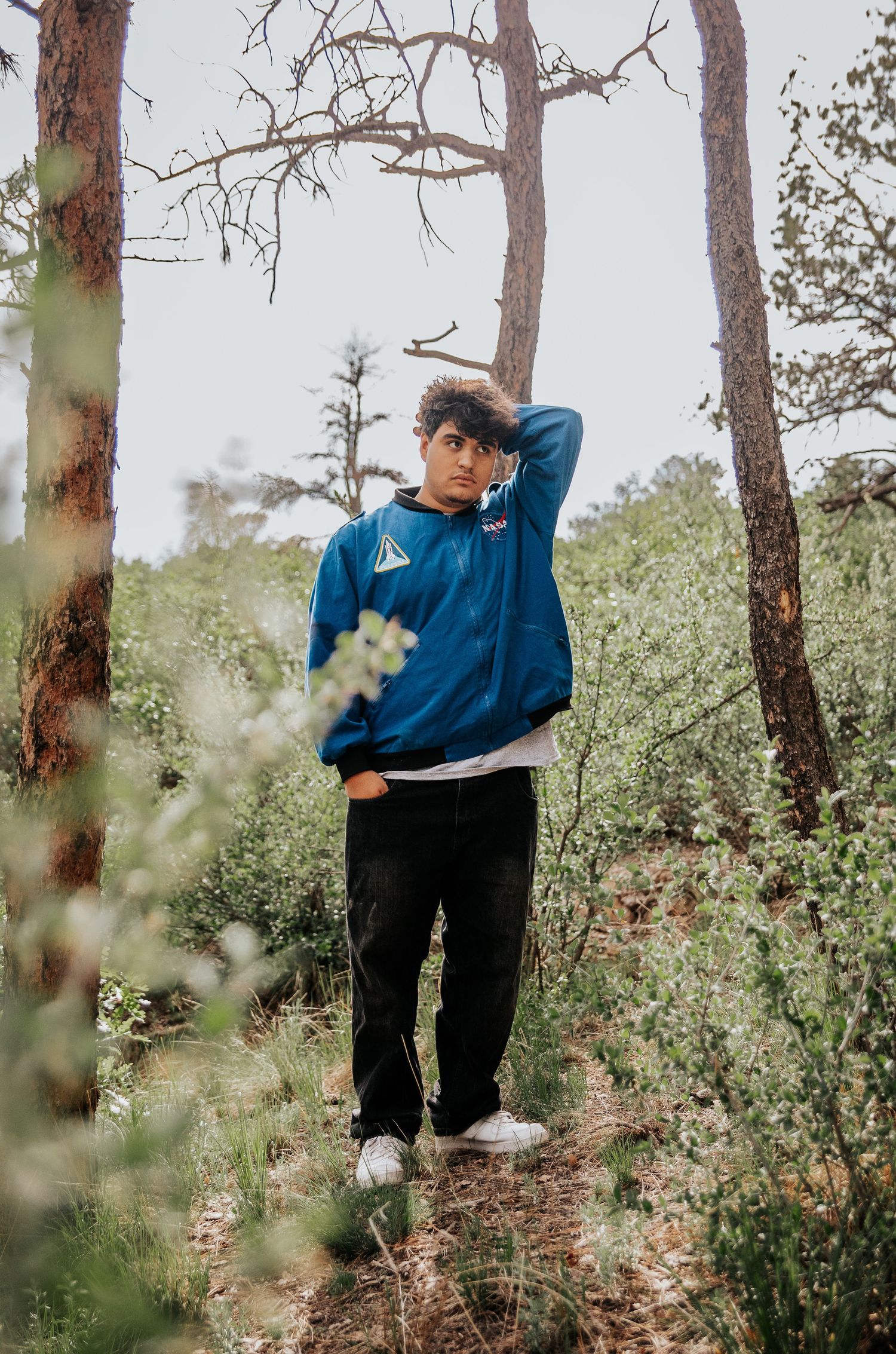 High school senior wearing a blue NASA jacket poses among tall pine trees in a wooded area at Palmer Park in Colorado Springs