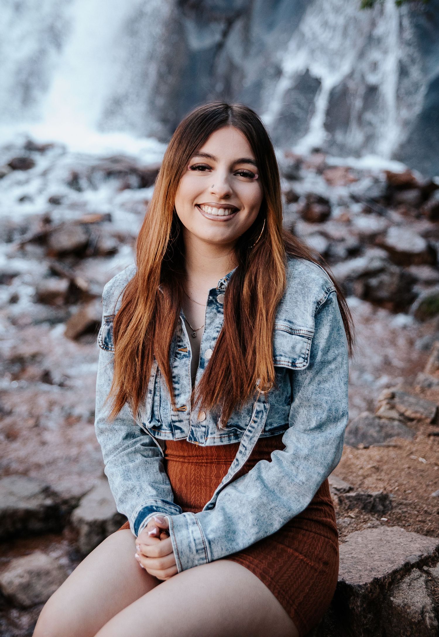 Portrait by a cascading waterfall wearing denim jacket and brown dress on rocky terrain.