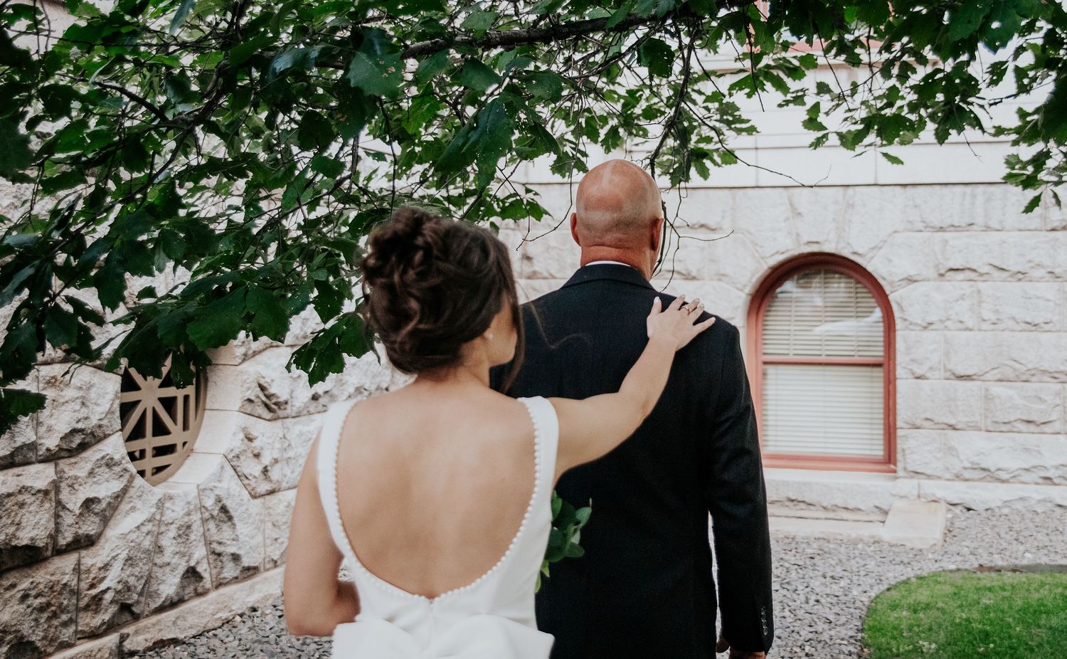 A couple in wedding attire stands together outside a white stone church building under green leaves.