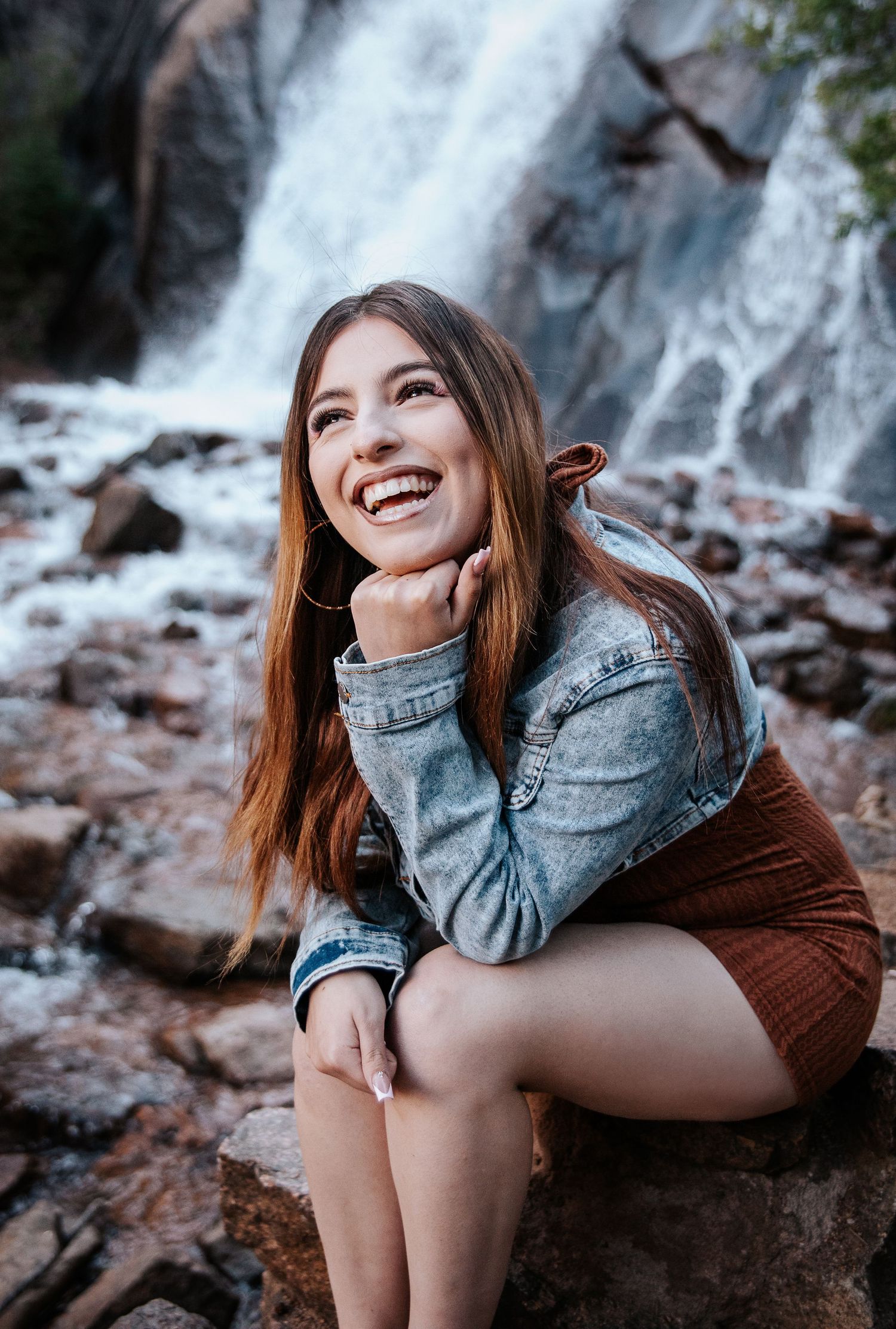 Casual outdoor portrait against waterfall backdrop with denim jacket and brown outfit.