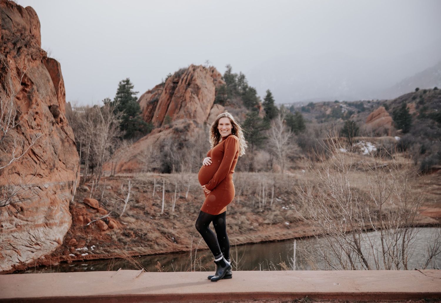 An expecting couple poses against dramatic red rock formations during golden hour.