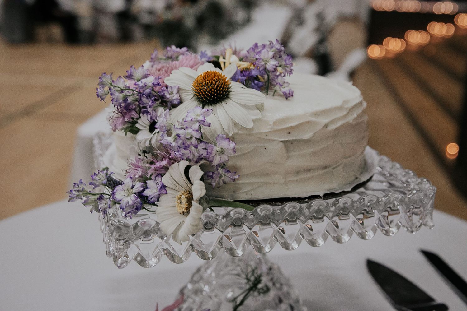 White wedding cake decorated with purple lilacs and a sunflower on a crystal cake stand.