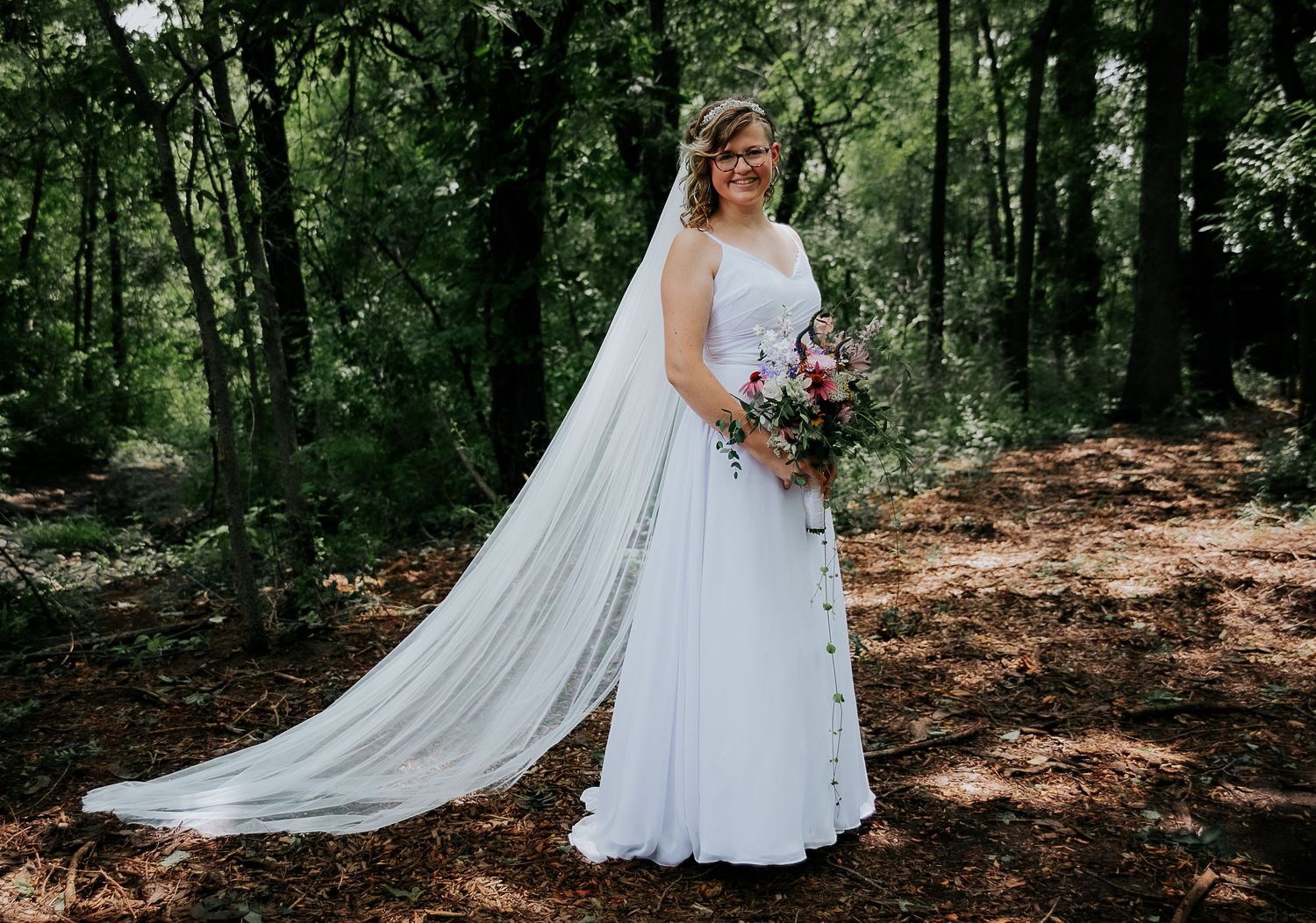 Bride in flowing white gown with long veil poses with bouquet in forest setting.