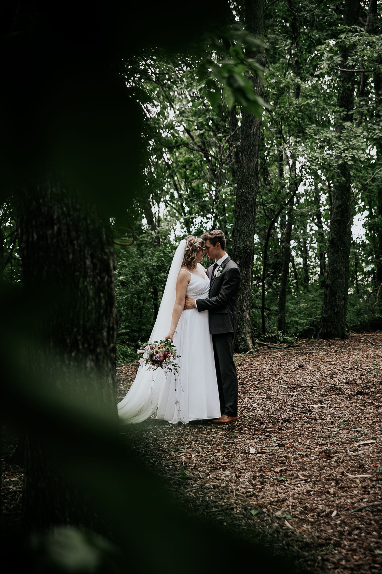 A wedding couple shares an intimate moment on a forest path surrounded by trees and fallen leaves.