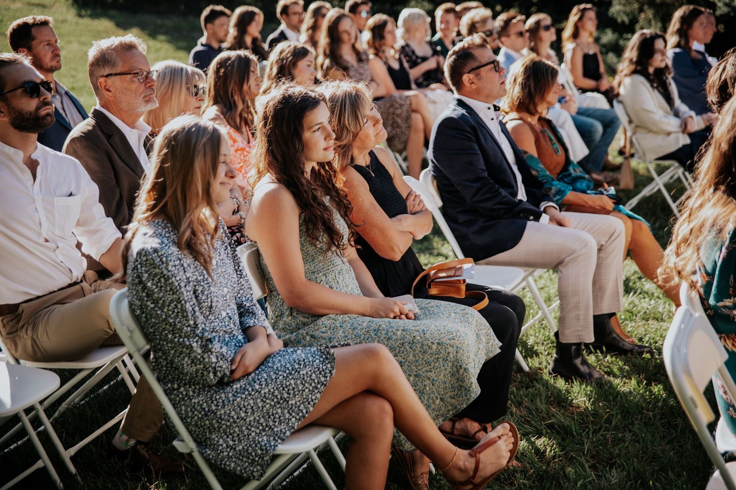 Wedding guests seated in white folding chairs watch an outdoor ceremony in late afternoon sunshine.