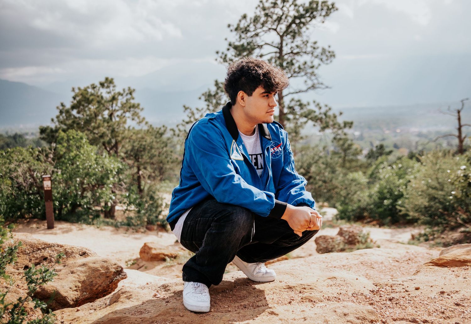 High school senior in NASA jacket sits on rocky ground along mountain hiking trail at Palmer Park in Colorado Springs