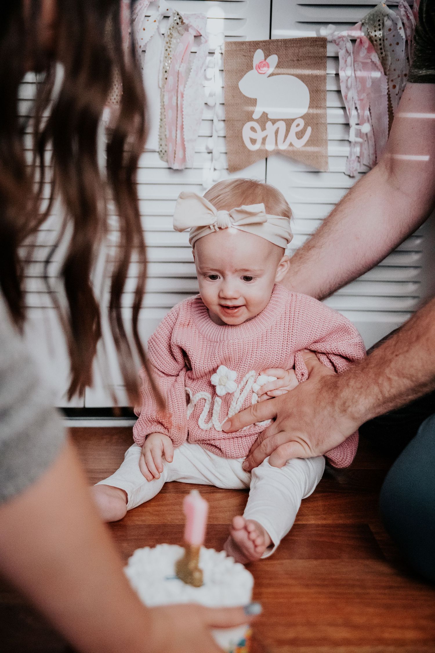 A cake smash photo session showing a small baby in a pink sweater reaching for a white frosted cake.
