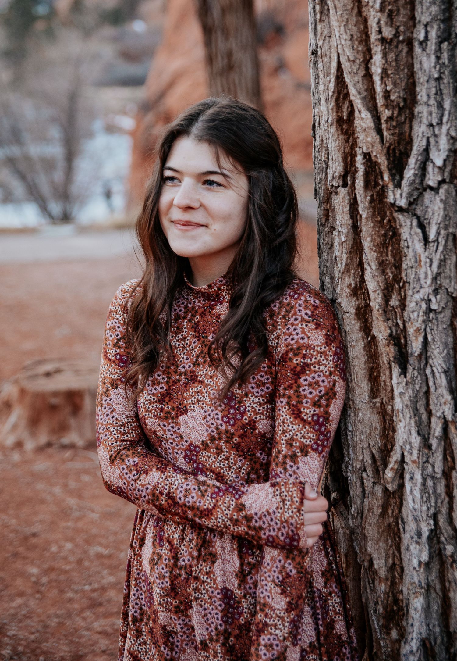 A woman in a rustic red floral dress poses against a tree trunk in a desert landscape.