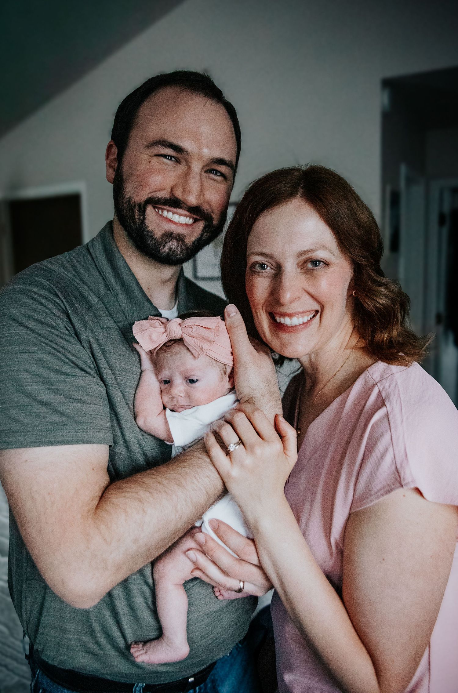 Happy couple holding their newborn baby wearing a pink bow headband in an indoor setting.